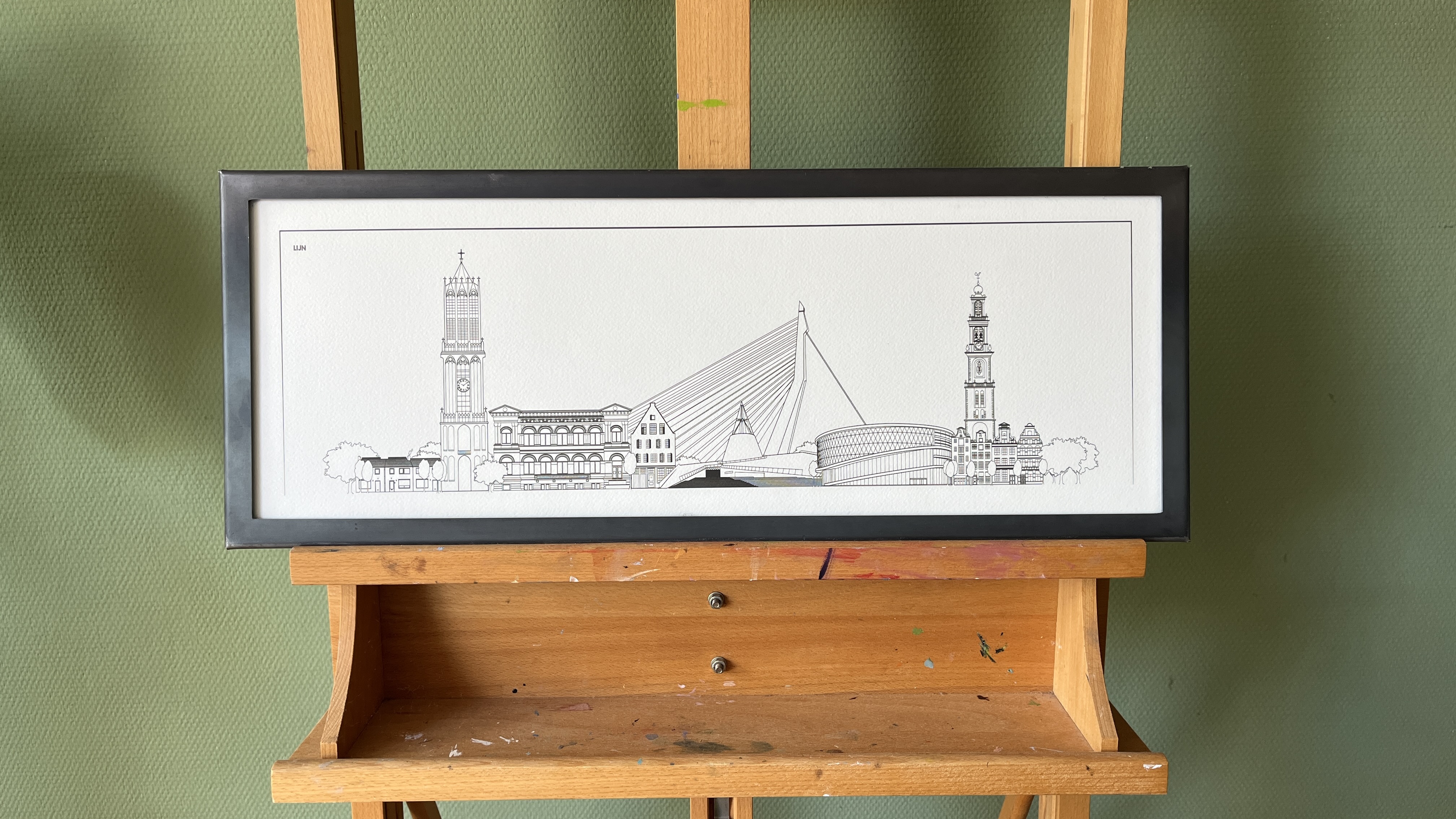 Ingelijste Line met een stadsgezicht: kerktoren, grachtenpand-achtige gevels en een kabelbrug, getoond op een schildersezel